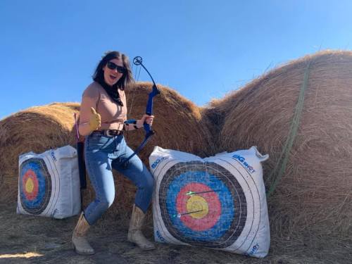 A woman poses with a bow and arrow beside hay bales and target boards, smiling confidently in a sunny outdoor setting.