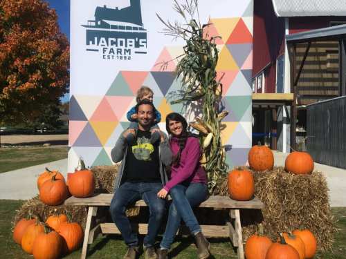 A family poses on a bench surrounded by pumpkins, with colorful autumn leaves and a farm backdrop.