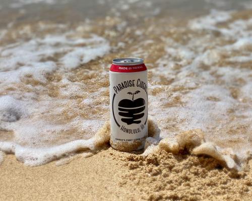 A can of Paradise Cider sits on sandy beach shore, partially covered by gentle ocean waves.