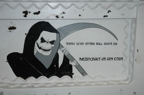 A grim reaper figure holding a scythe, with the text: "Those who oppose will meet me. Democracy at any cost."
