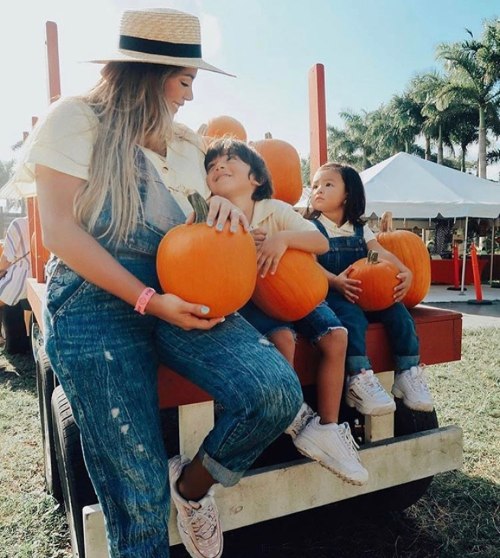 A woman and two children sit on a wagon, each holding pumpkins, with palm trees and a blue sky in the background.
