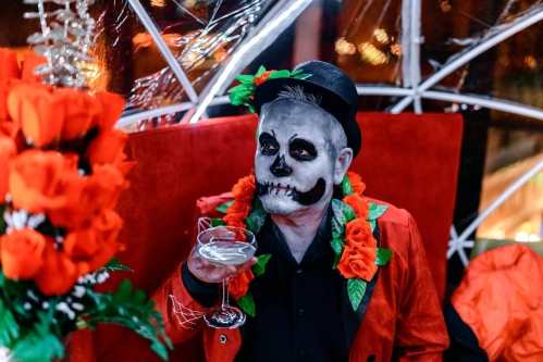 A person in a skeleton costume with face paint, wearing a hat and floral decorations, holding a cocktail in a festive setting.
