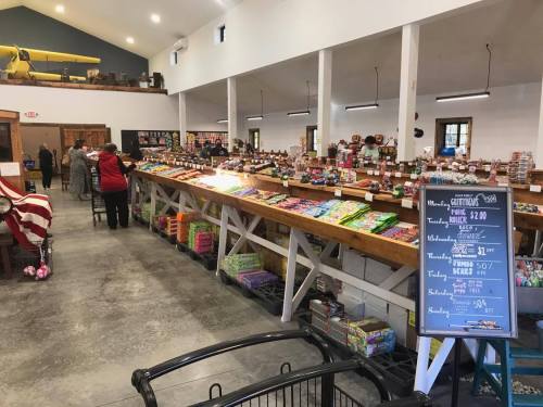 A spacious market interior with shelves of colorful candies and snacks, and a chalkboard menu displaying prices.