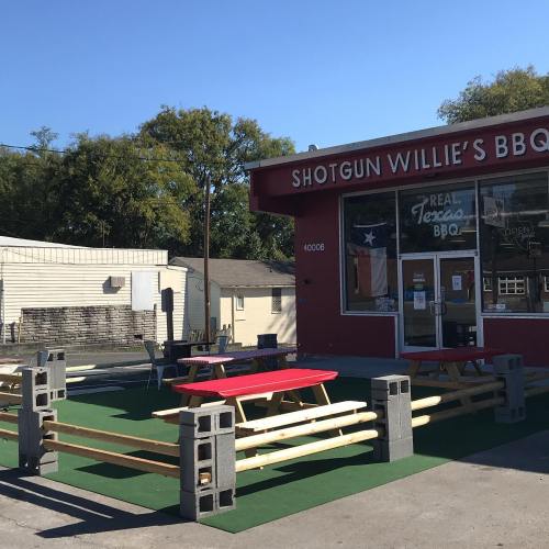 Outdoor seating area with red picnic tables and benches in front of Shotgun Willie's BBQ restaurant. Clear blue sky.