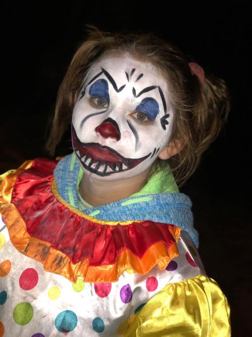 A child dressed as a clown with colorful makeup and a polka dot costume, smiling in a dark setting.