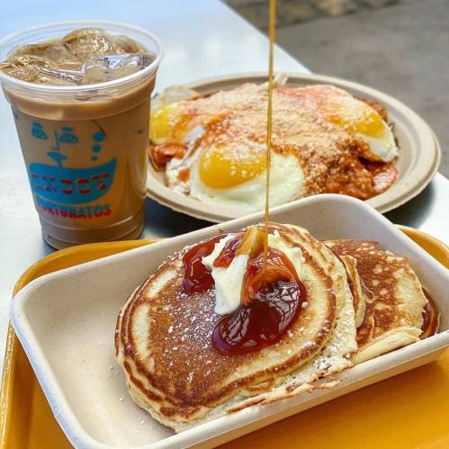 A plate of pancakes with butter and syrup, alongside a dish of eggs and a cup of iced coffee.