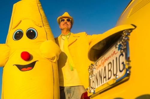 A person in a yellow suit and sunglasses stands next to a large yellow banana costume by a car with a playful license plate.
