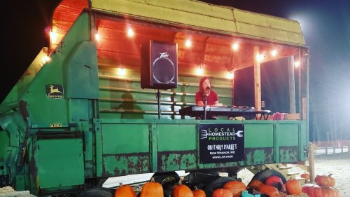 A person performs on a stage made from a green tractor trailer, surrounded by pumpkins and string lights at night.