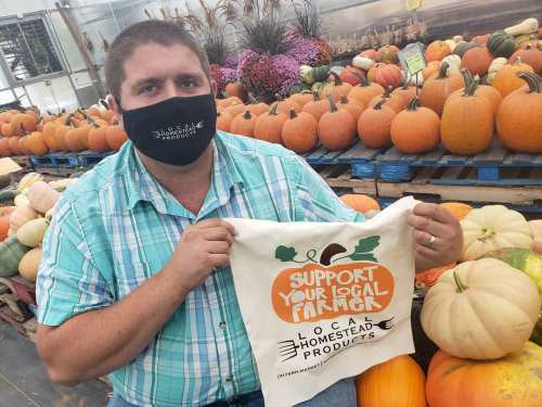 A farmer in a mask holds a tote bag that says "Support Your Local Farmers" among various pumpkins.