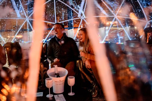 A couple laughs together inside a cozy, illuminated dome, enjoying drinks amidst a festive atmosphere.