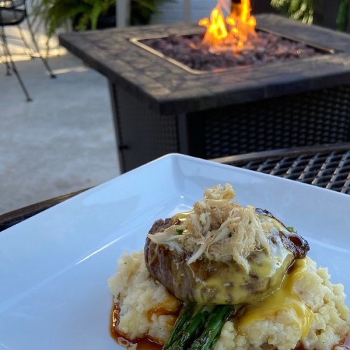 A gourmet dish featuring a meat patty topped with cheese and shredded chicken, served on mashed potatoes with asparagus.