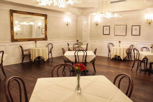 Elegant dining room with round tables, white tablecloths, and floral centerpieces, featuring chandeliers and a large mirror.