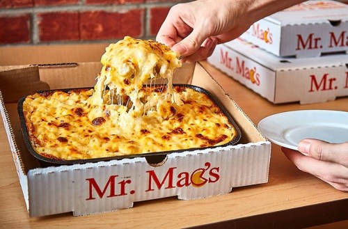 A hand lifts a cheesy, baked macaroni dish from a takeout box labeled "Mr. Mac's," with more boxes in the background.