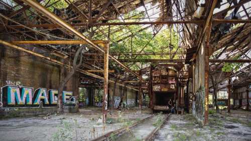 Abandoned industrial space with rusted metal beams, overgrown vegetation, and graffiti on the walls.