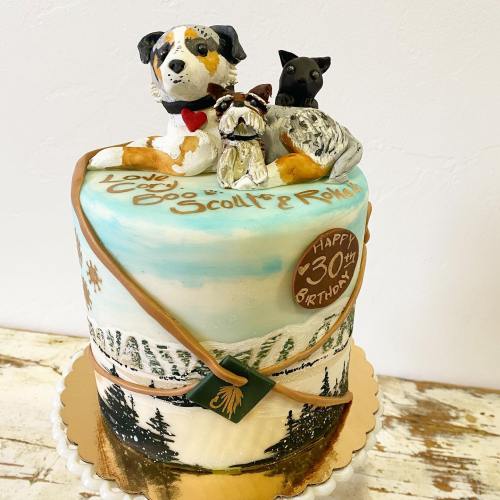 A beautifully decorated birthday cake featuring three dog figurines and a scenic landscape design.