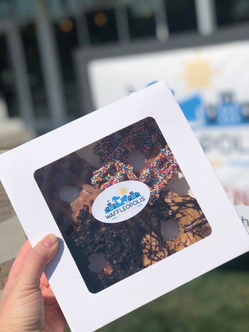 A hand holds a white box with a clear window displaying colorful, decorated waffles from Waffledopolis.
