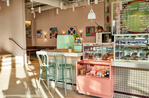 A bright café interior featuring pastel decor, a display case of treats, and colorful artwork on the walls.
