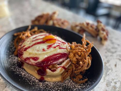 A plate of pancakes topped with a sunny-side-up egg, berry sauce, and crispy onion strings, with waffles in the background.