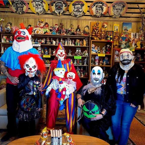 A group of people in clown masks poses with clown dolls in a colorful room filled with memorabilia.