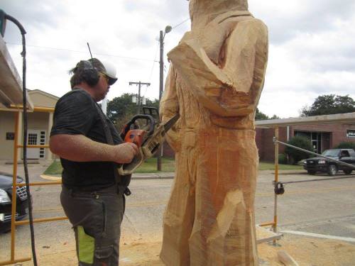 A man uses a chainsaw to carve a wooden statue, surrounded by wood shavings and scaffolding.