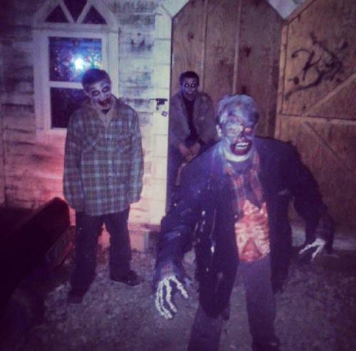 Three zombies in tattered clothing stand outside a boarded-up building, with eerie lighting enhancing their spooky appearance.