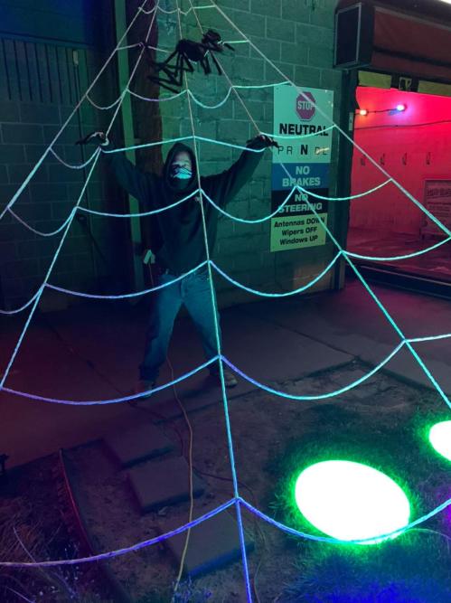 A person in a hoodie stands in a colorful spider web display, with a large spider above and glowing lights below.