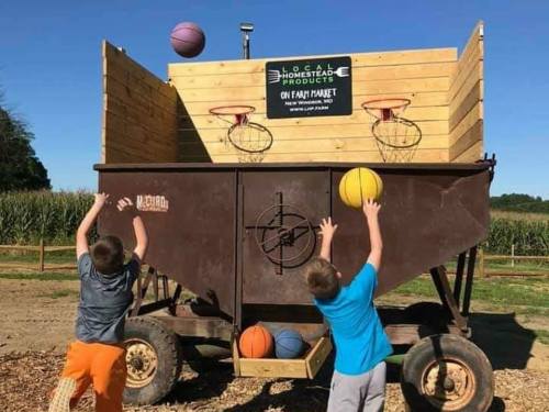 Two children throw basketballs at a makeshift hoop on a trailer in a sunny outdoor setting.