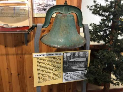 A vintage school bell displayed with an informational sign about Quosatne/Hawkins School, set against a wooden background.