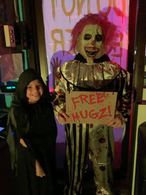 A child in a black cloak stands next to a clown in a bloody costume holding a sign that says "FREE HUGZ!"
