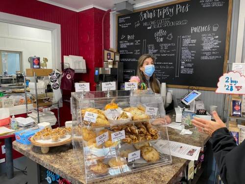 A woman in a mask stands behind a counter displaying various baked goods in a cozy café setting.