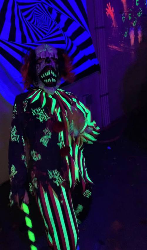 A creepy clown in a brightly colored, striped costume glows under blacklight, surrounded by spooky decorations.