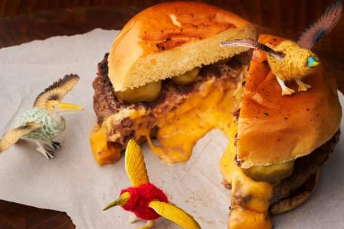 A cheeseburger with melted cheese and pickles, partially cut, surrounded by colorful toy birds.