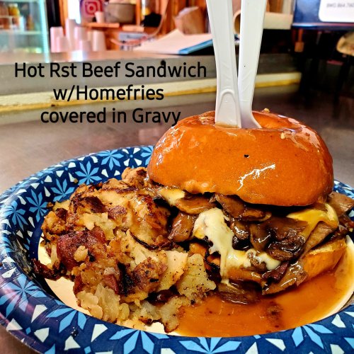 A hearty roast beef sandwich topped with melted cheese, served with home fries and gravy on a blue paper plate.