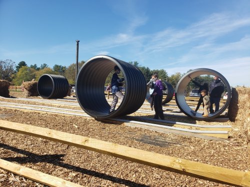 Children and adults play on large, cylindrical tubes in a sunny outdoor setting with wooden paths and mulch.