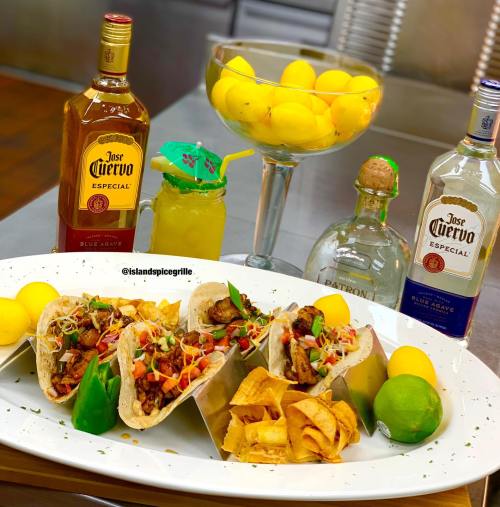 A vibrant table setting featuring tacos, chips, and drinks, with bottles of tequila and a bowl of lemons in the background.