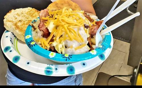 A person holds a plate with a biscuit and a bowl of loaded breakfast with eggs, bacon, cheese, and gravy.