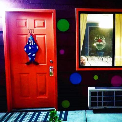 A bright orange door with a clown decoration, next to a window showing a surprised face and a clown painting inside.