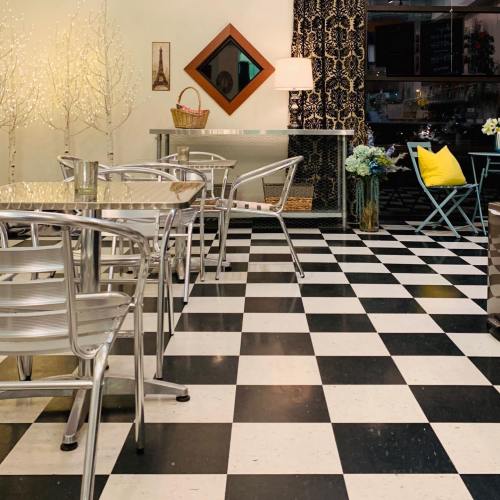 A cozy café interior with a checkered floor, metal chairs, decorative trees, and a stylish mirror on the wall.