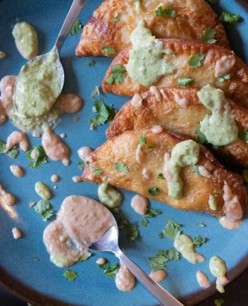 Four golden-brown empanadas on a blue plate, drizzled with green and pink sauces, garnished with fresh cilantro.
