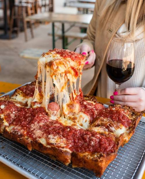 A person holds a glass of wine while pulling a slice of cheesy, saucy pizza from a large tray.