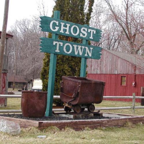 Sign for "Ghost Town" with old mining carts in front, set in a rural area with wooden buildings in the background.
