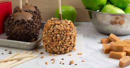 A caramel apple coated in chopped nuts, alongside chocolate-covered apples and green apples in a bowl.