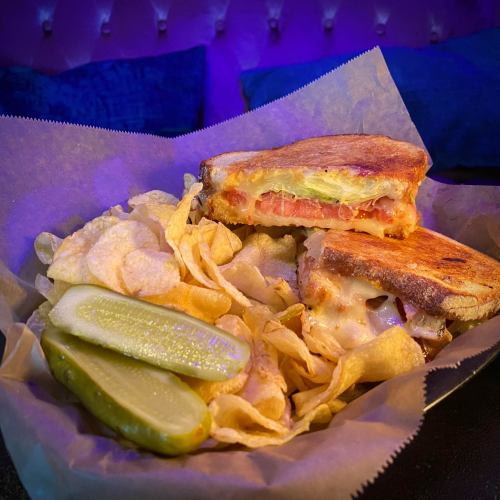 A grilled sandwich with melted cheese and tomatoes, served with potato chips and pickles on a parchment-lined plate.