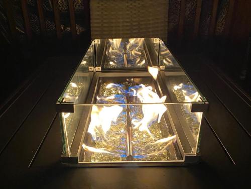 A modern fire pit with glass sides, featuring flames dancing over decorative glass stones in a dark setting.
