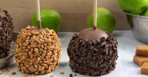 Two caramel apples coated in chopped nuts and chocolate shavings, with green apples and caramel pieces in the background.