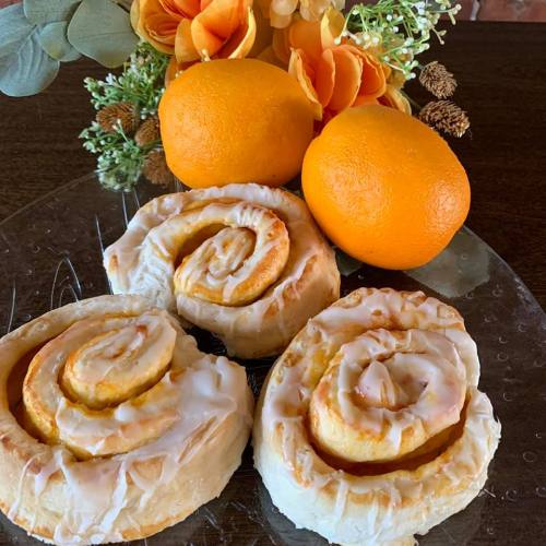 Three orange cinnamon rolls with icing, accompanied by fresh oranges and a bouquet of flowers on a wooden table.