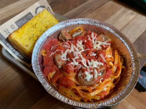 A foil container with fettuccine pasta in marinara sauce, topped with cheese and sausage, alongside a piece of garlic bread.