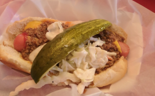 A hot dog topped with chili, cheese, shredded lettuce, and a pickle slice, served in a soft bun.