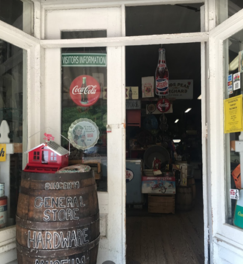 Entrance to a vintage general store with a Coca-Cola sign, wooden barrel, and various items visible inside.