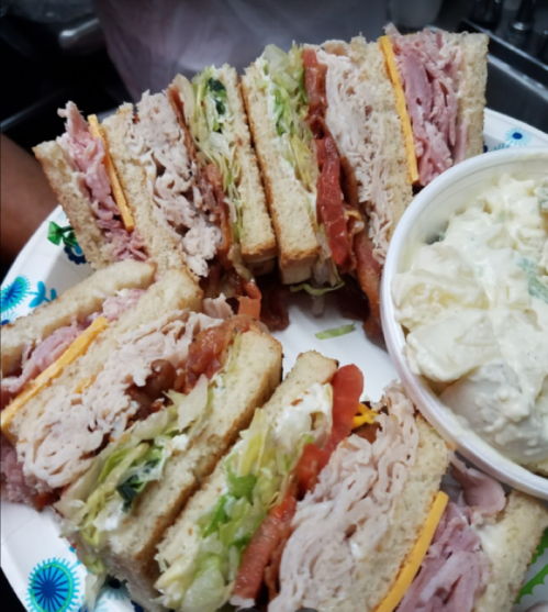 A platter of stacked sandwiches with turkey, ham, cheese, lettuce, and tomato, alongside a bowl of potato salad.